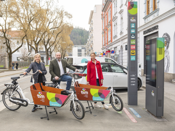 Drei Personen mit tim-Lastenrädern und E-Auto in Graz. Infosäule im Hintergrund.